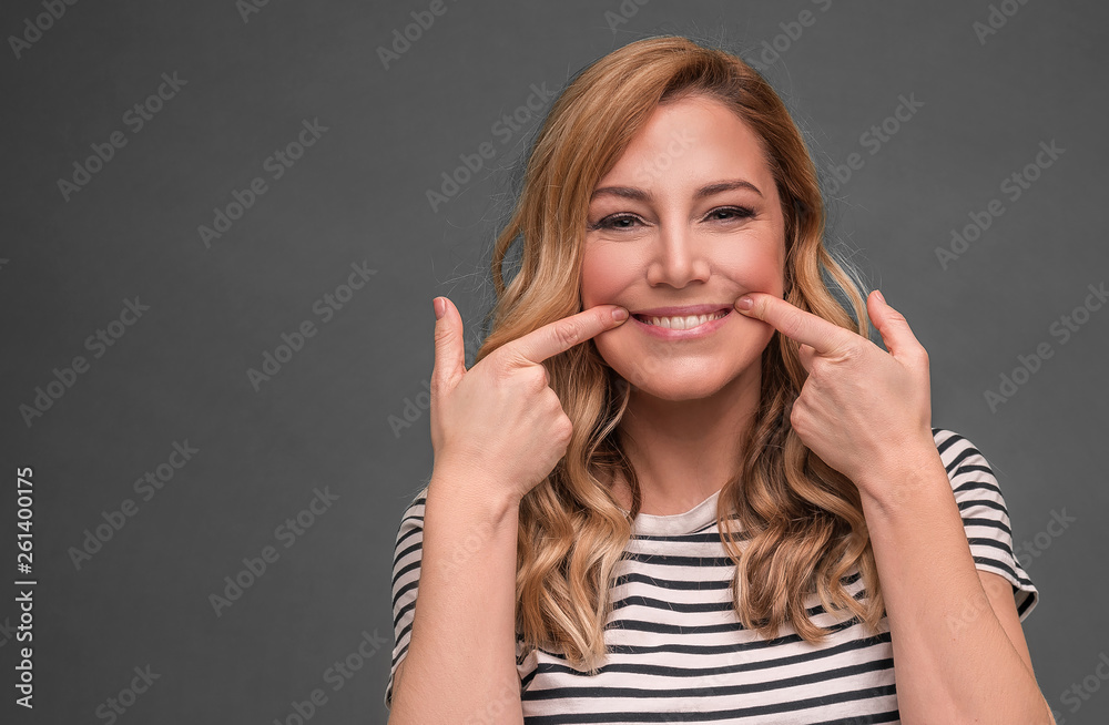 Fototapeta premium A young woman stretches her mouth in an insincere smile against a gray background. Fake smile.