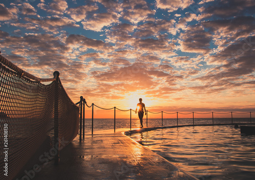 Swimmer wlaking along the side of Bronte Beach Pool at Sunrise with sunburst. Epic cloudscape.