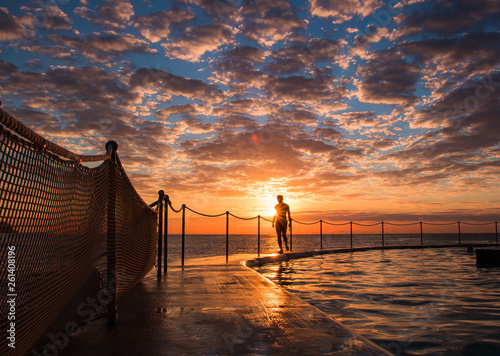 Swimmer walking along the side of Bronte Beach Pool at Sunrise with sunburst. Epic cloudscape.