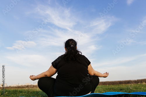 Body positive, yoga, meditation, tranquility, relax. Overweight woman meditating sitting at yoga mat in the meadow