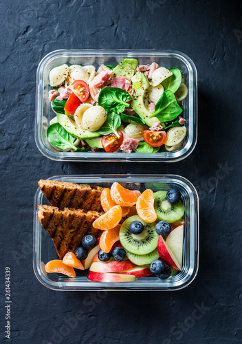 Office sweet and savory food lunch box. Pasta, tuna, spinach, avocado salad and fruit, peanut butter sandwiches lunch box on dark background top view