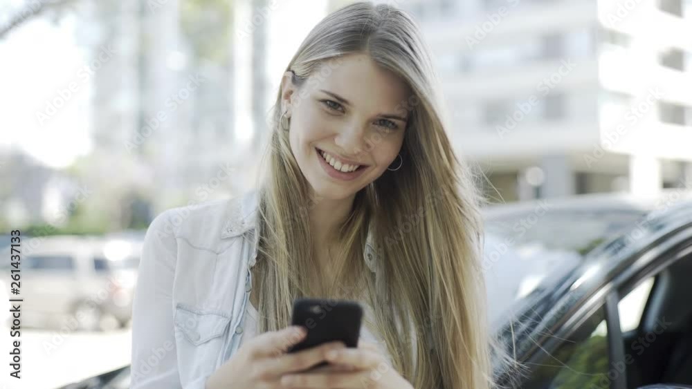 Medium shot of smiling young woman using smartphone while standing near car Medium shot of smiling young woman using smartphone while standing near car