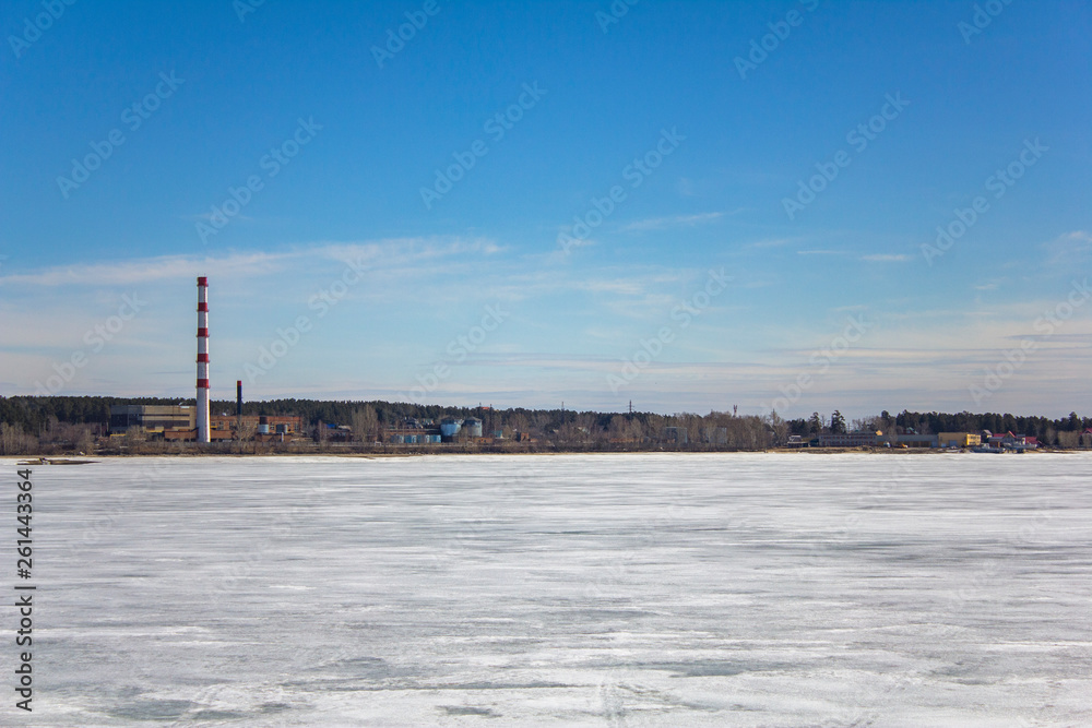 frozen white snow lake on the background of a plant with a striped red pipe, buildings and warehouses in a green forest under a blue sky