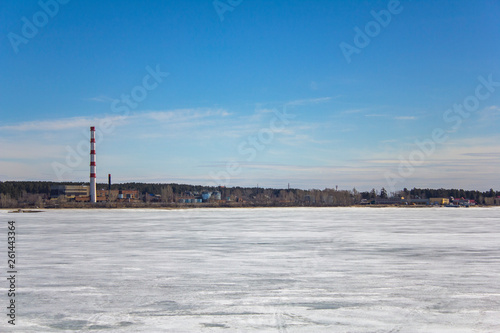 frozen white snow lake on the background of a plant with a striped red pipe, buildings and warehouses in a green forest under a blue sky © Pavel