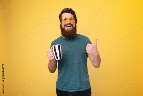 Photo of bearded man in stylish eyeglases, hooldding cup and doing like gesture over yellow background photo