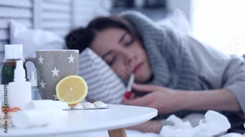 Beautiful sick ill woman female adult with scarf lying on bed with flu at grey bedroom with medicine, drugs and cup of tea with lemon, coughing and measuring temperature by electric thermometer.