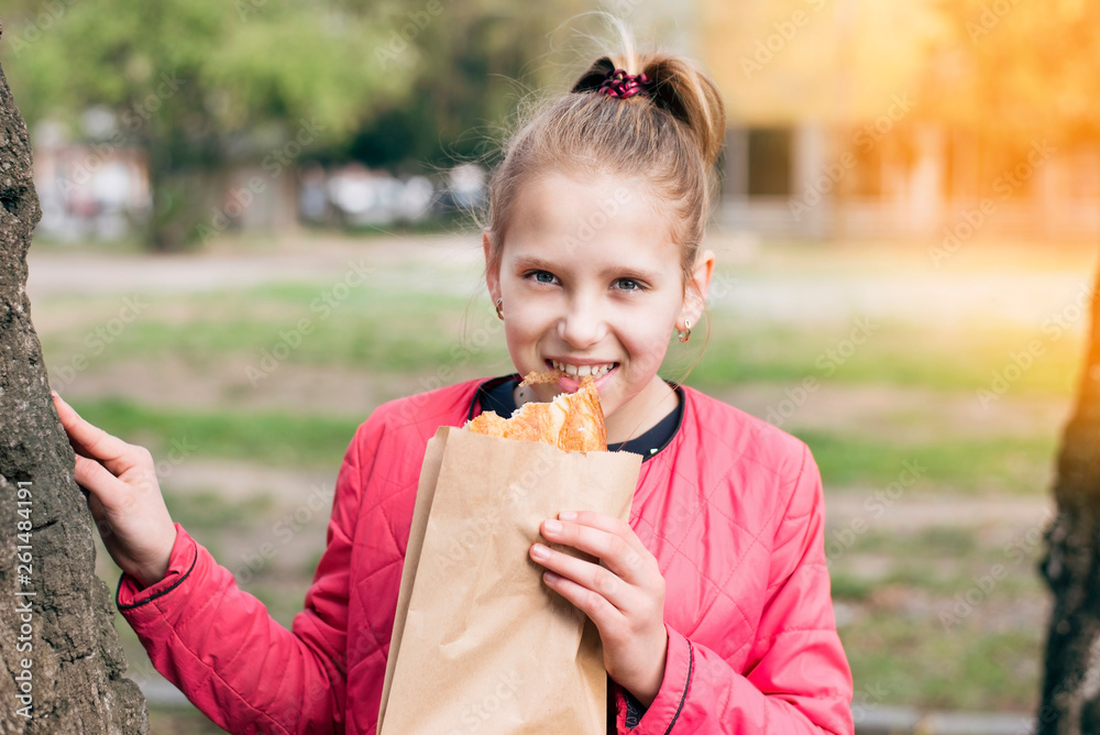 A beautiful young girl holds a paper bag in her hands and eats a bun that sale in the paper package A beautiful young girl holds a paper bag in her hands and eats a bun that sale in the paper package