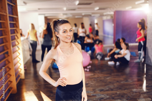 Fototapeta Naklejka Na Ścianę i Meble -  Young smiling Caucasian brunette in sportswear holding hand on hip, posing and looking at camera. In background women resting after exercises. Gym interior.