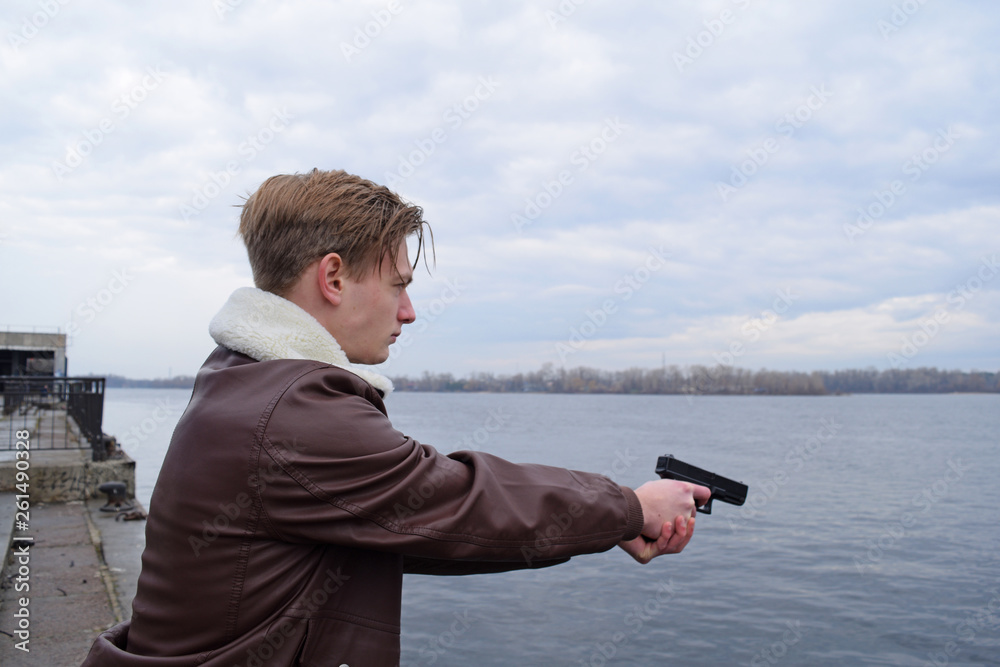 A young guy in a leather jacket shoots with a gun near the river. A young guy in a leather jacket shoots with a gun near the river.