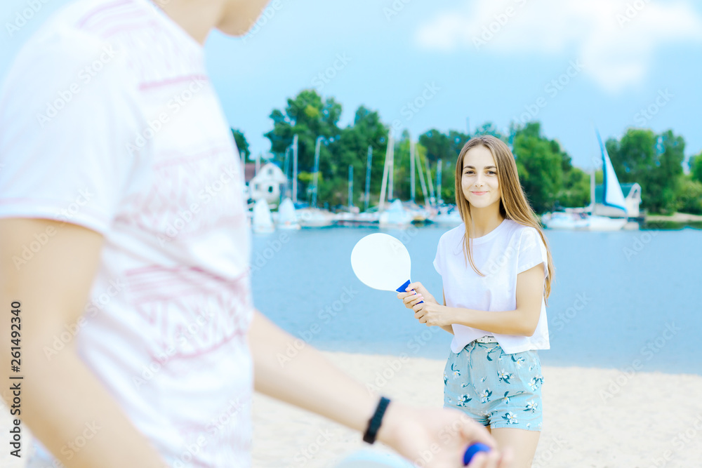 Male is holding blue ball, his smiling girlfriend ready to catch with racket on background. Young man and woman in light clothes are playing in matkot on beach. Summer activities concept. Male is holding blue ball, his smiling girlfriend ready to catch with racket on background. Young man and woman in light clothes are playing in matkot on beach. Summer activities concept.