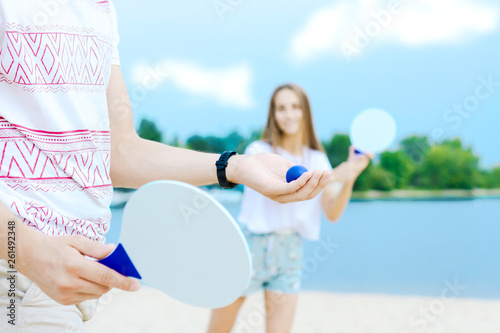 Closeup male hands holding white racket, his smiling girlfriend ready to catch on background. Young man and woman in light clothes are playing in matkot on beach. Summer activities concept.