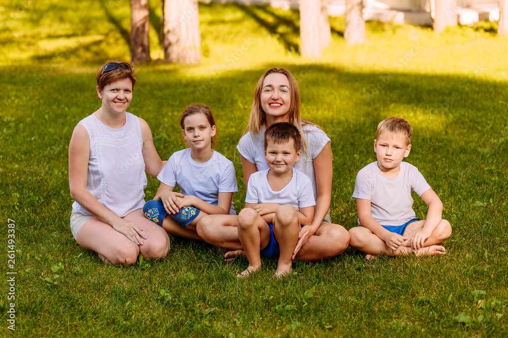 smiling mothers and children sit on the green grass. Big happy family, two women and three children in white t-shirts smiling mothers and children sit on the green grass. Big happy family, two women and three children in white t-shirts