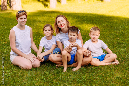 smiling women and children sit on the green grass. Big happy family, two mothers and three children in white t-shirts. Summer © Alyona