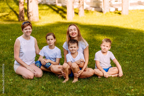happy women and children sit on the green grass and look at the camera. Big happy family, two mothers and three children in white t-shirts and shorts. © Alyona