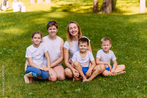 Two happy young mothers and three children in white t-shirts sit on a green lawn in the summer. Big Caucasian family on vacation. Women hug children.