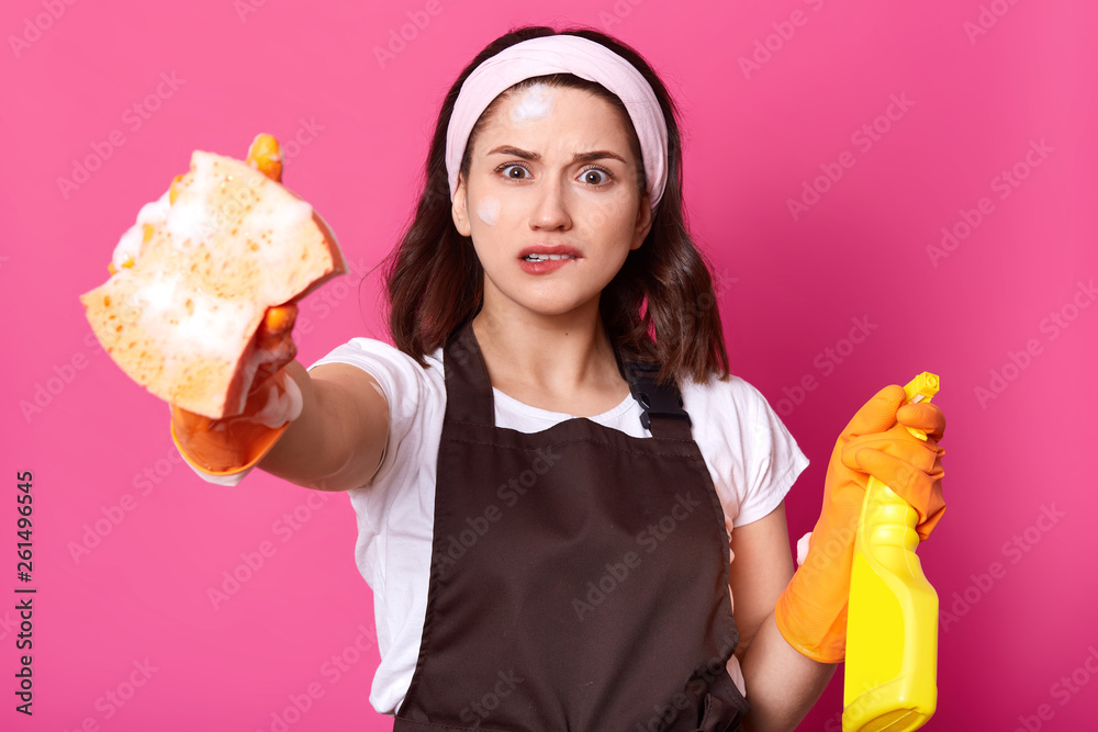 Frustrated young brunette female in brown apron and orange protective gloves, holds sponge and detergent, biting her lip, feeling stressed, Housekeeping, housework, hygiene and cleaniness concept. Frustrated young brunette female in brown apron and orange protective gloves, holds sponge and detergent, biting her lip, feeling stressed, Housekeeping, housework, hygiene and cleaniness concept.