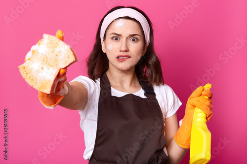 Frustrated young brunette female in brown apron and orange protective gloves, holds sponge and detergent, biting her lip, feeling stressed, Housekeeping, housework, hygiene and cleaniness concept. photo