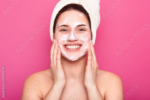 Studio shot of charming young woman, over rose background, with white towel on her head and cream on her face, posing with toothy smile, touches her cheeks with hands, looks happy and satisfied.