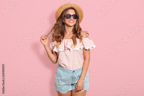 Beautiful young woman in rose summer blouse, blue short, sunglasses and sun hat, pulling her hair aside, being in good mood, ready for going out with friends. Studio shot over pink background.