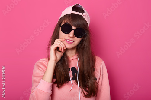 Studio shot of attractive brunette pretty teenager, wears hoodie and cap, has long dark hair, keeps fingers near mouth, poses on pink studio background, advertises clothes from new collection. © sementsova321