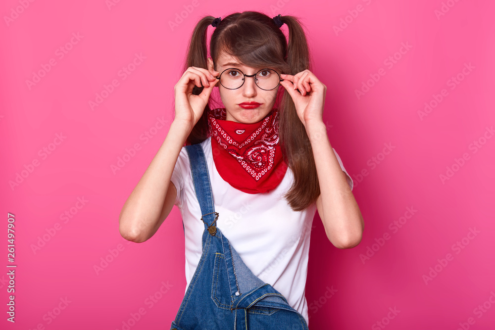 Disappointed charismatic young lady with bangs and long pigtails has frown face, touches her eyeglasses, choosing comfortable position. Youth, lifestyle and emotions concept. Copy space for ad.