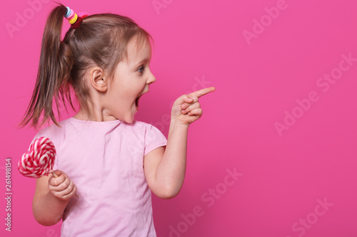 Close up portrait of little attractive child opening mouth widely, looking other side with excitement, holding heart bright lollipop. Playful merry little fair haired girl spends sparetime happily. photo
