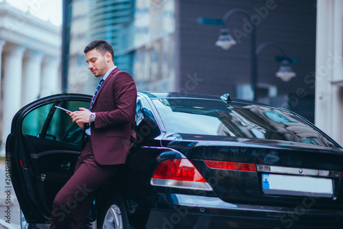 Handsome young businessman leaning on his limo and smiling while looking at his phone. © qunica.com