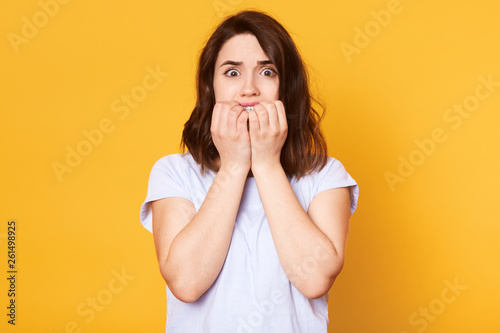 Studio shot of young brunette woman wearing casually, nervous and bites nails, standing with eyes full of fear, attractive female afraids of something, photo isolated on concentrated yellow background