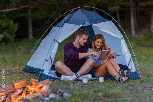 Young couple drinking coffee, enjoying the mountain view and checking the tablet with bonfire in the sunset