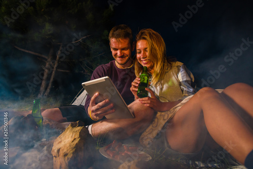 Young couple baking sausages on the campfire, drinking beer and enjoying the tablet pc in the forest hill in the dusk photo