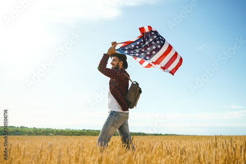 4th of July. Fourth of July. American with the national flag. American Flag. Independence Day. Patriotic holiday. The man is wearing a hat, a backpack, a shirt and jeans. Beautiful sunset light. 