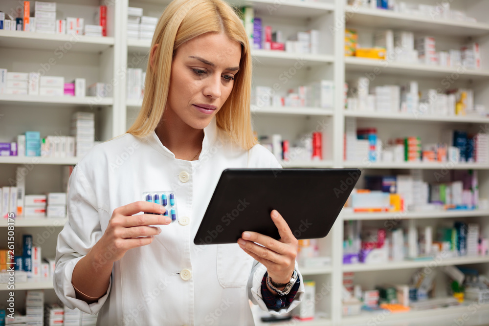 Serious young female pharmacist with blond hair using a tablet to check product description, holding pack of pills in other hand. Shelves with medications in the background