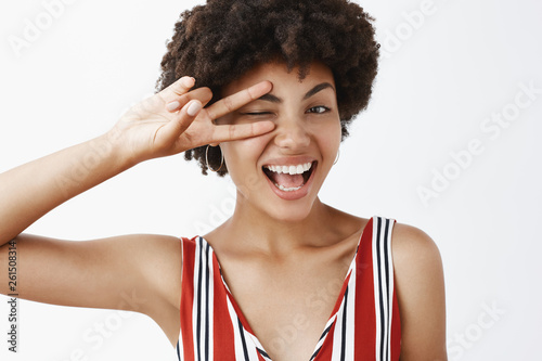 Pure joy and happiness. Portrait of satisfied happy and feminine dark-skinned woman feeling amused and excited  greeting best friends  winking and smiling broadly  showing victory sign over eye
