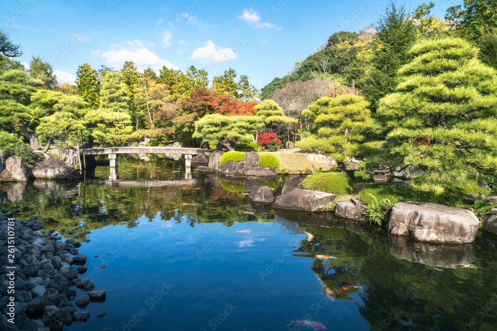 Small stone bridge over the pond, full of Koi fish, at Koko-en Garden on a beautiful sunny day, with the rooftop of Himeji Castle just peeking over the tree line, in Japan.