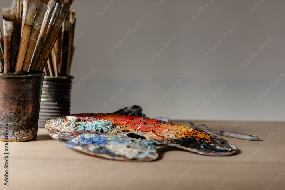 Fototapeta premium Artist's studio - jar of paint brushes on a wooden table and a colorful palette with paint on