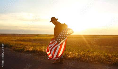 4th of July. Fourth of July. American with the national flag. American Flag. Independence Day. Patriotic holiday. The man is wearing a hat  a backpack  a shirt and jeans. Beautiful sunset light. 