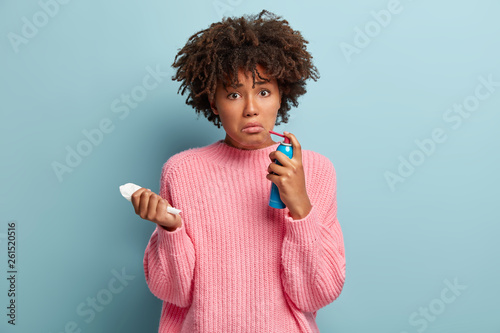 People, treatment and disease concept. Discontent Afro American female cures sore throat with spray, suffers from cough and bronchitis, holds white handkerchief, frowns face, wears pink jumper