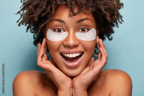 Bilde på lerret Pure beautiful woman takes care of skin, has under eye patches, keeps hands on face, smiles broadly, has white teeth, Afro haircut, models over blue background