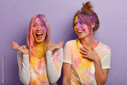Indian holidays, fun, festival concept. Cheerful joyful women play with colors, smeared with multicolored powder, participate in Holi fest which denotes triumph of good over evil, laugh, pose indoor © Wayhome Studio