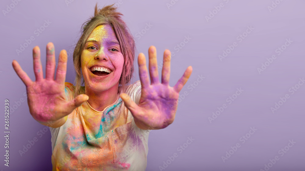 Indoor shot of glad positive Caucasian woman shows colorful palms, stretches hands, splashes powder, smiles positively, isolated over purple background with empty space. Holi festival concept - obrazy, fototapety, plakaty 