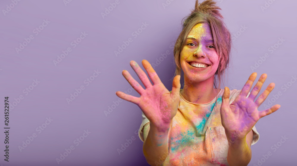 Happy Holi day concept. Funny satisfied girl with colorful powder on face,  clothes and palms, being in high spirit, celebrates traditional spring  festival, poses against purple wall with free space Stock Photo |