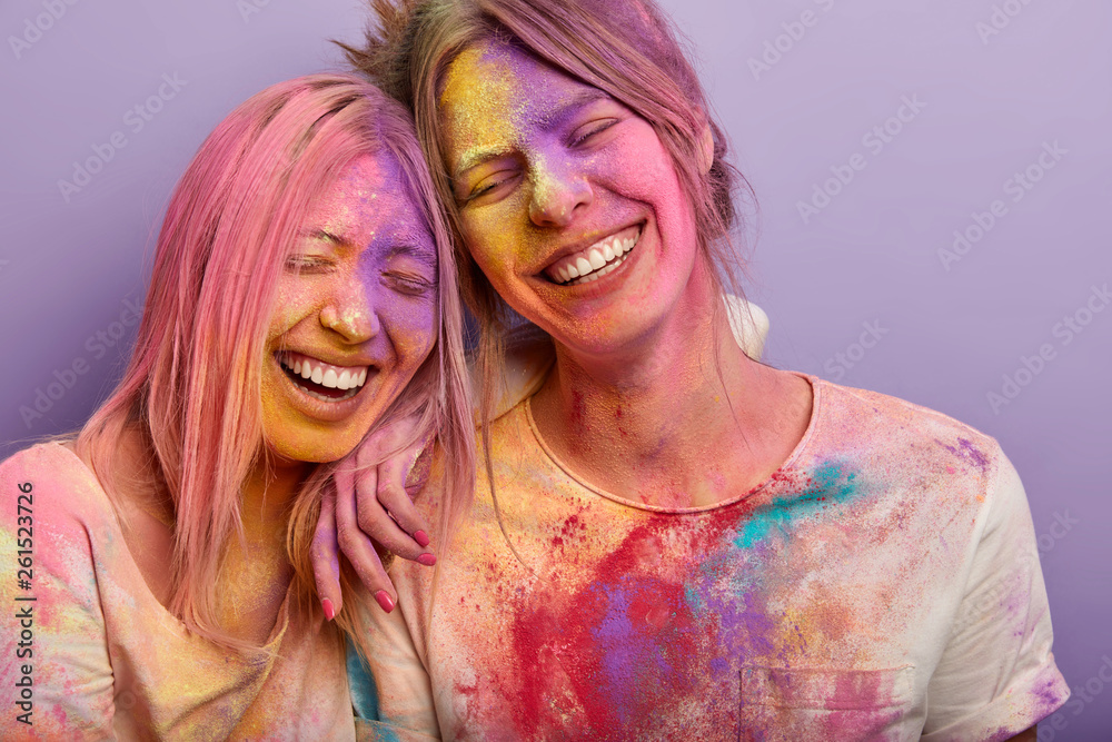 Sincere emotions and feelings concept. Funny two female friends lean heads  to each other, laugh gladfully, have broad smiles, colored dirty faces,  splattered clothes, participate in Holi festival Stock Photo | Adobe