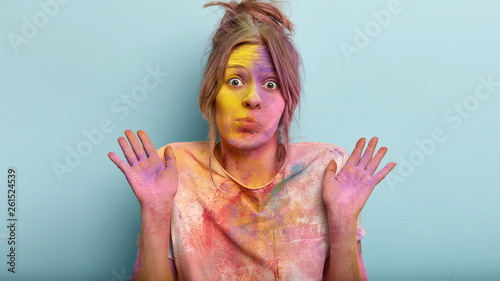 Indignant puzzled female raises hands, says she is not guilty, powdered with colorful dye, has confused facial expression stands over blue background dirty after playing with colors on spring festival