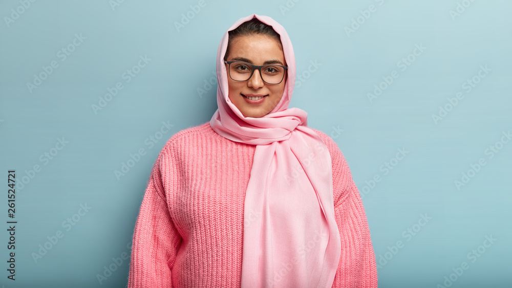 Half length shot of beautiful dark eyed smiling female in pink jumper and hijab, looks through spectacles, happy to hear good news, follows Muslim religion and rules, has satisfied gaze in camera Half length shot of beautiful dark eyed smiling female in pink jumper and hijab, looks through spectacles, happy to hear good news, follows Muslim religion and rules, has satisfied gaze in camera
