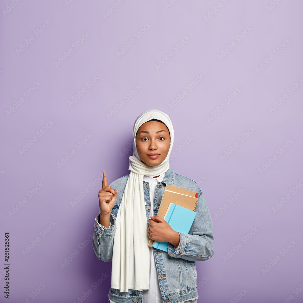 Surprised intriguing woman in headwear points upwards and looks with interest, shows blank space above for your advertisement or information, carries diary and spiral notebook. Muslim religion Surprised intriguing woman in headwear points upwards and looks with interest, shows blank space above for your advertisement or information, carries diary and spiral notebook. Muslim religion