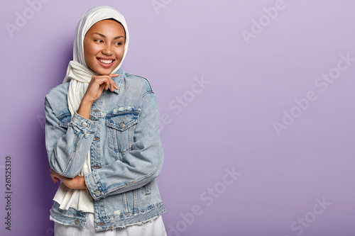 Religious Arabic woman has cheerful expression, covers head with white hijab, wears denim jacket, holds chin, looks away, stands against purple background. People, ethnicity and faith concept