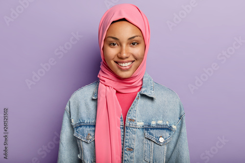 Studio shot of glad Eastern female has Islamic religion, covered head with pink veil, smiles gently, shows white teeth, isolated against violet wall expresses positive feelings and emotions. Ethnicity