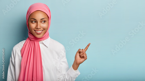 Smiling black woman has cheerful expression, points away with fore finger, shows blank space on right corner, has modest look, wrapped in pink veil, isolated over blue background. Look at this photo