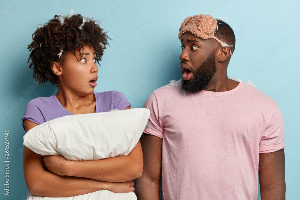 Embarrassed scared dark skinned woman and man look at each other with shock, wear nightwear, eyemask dissatisfied by bad sleep, surprised to hear loud music from neighbours, isolated over blue wall Embarrassed scared dark skinned woman and man look at each other with shock, wear nightwear, eyemask dissatisfied by bad sleep, surprised to hear loud music from neighbours, isolated over blue wall
