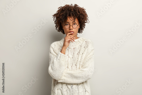 Photo of thoughtful dark skinned female has curly hair, serious look, being deep in thoughts, looks straightly at camera, wears long sleeve sweater, isolated over white background. Thinking concept